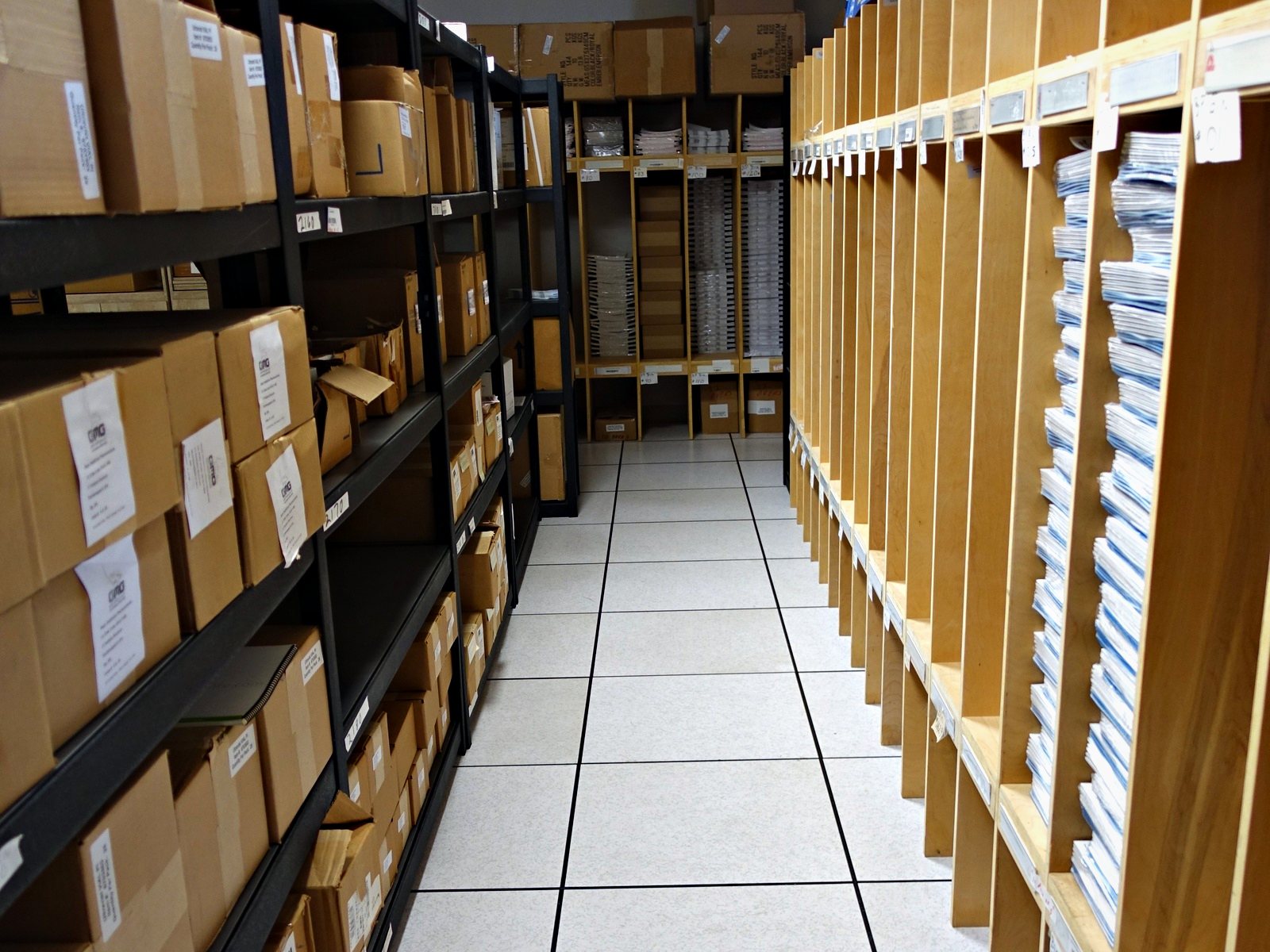 An aisle of storage at Broudy Printing: labelled cartons on one side, printed work in pigeonholes on the other